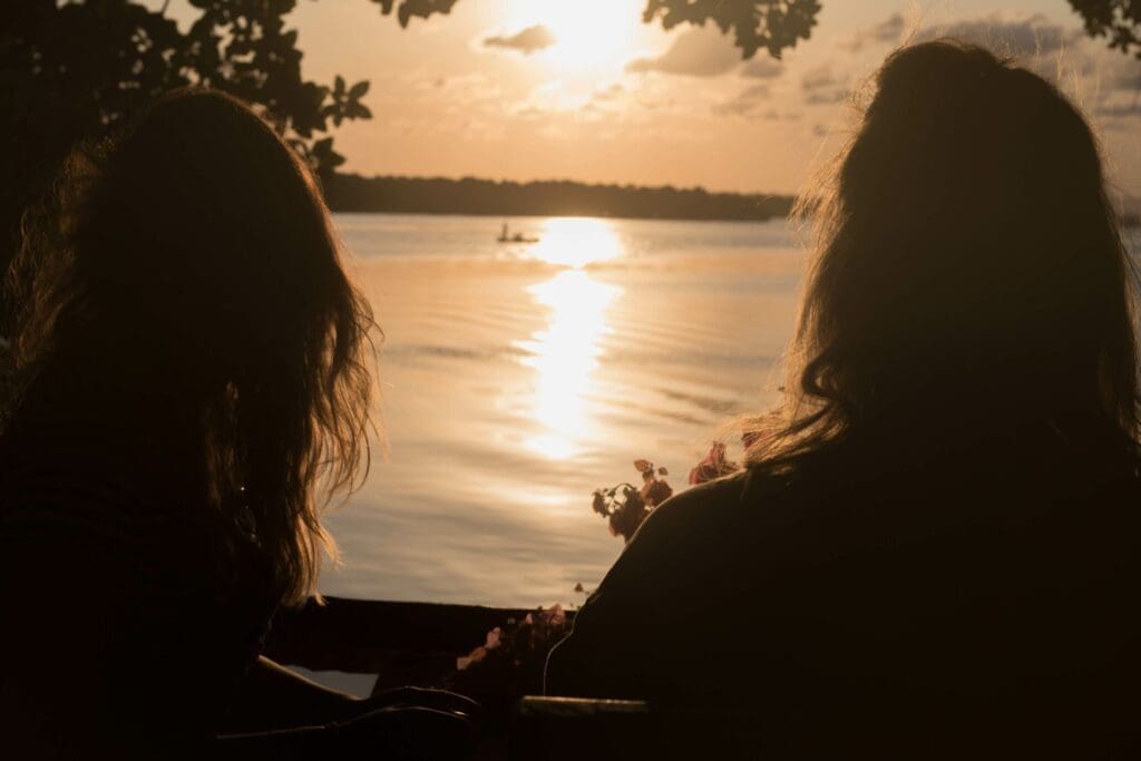 Twee mensen zitten aan een rustig meer bij zonsondergang, hun gezichten in de schaduw terwijl ze naar het water staren dat de gouden zon weerspiegelt, met boomtakken die het tafereel omlijsten en een kleine boot in de verte.