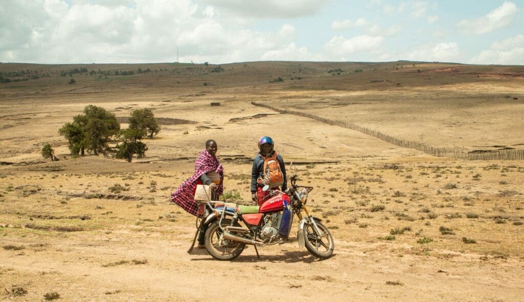 A person in traditional Maasai clothing stands beside a motorcycle with a rider in a blue helmet and orange jacket, set against Kenya’s vast, dry, open landscape under a partly cloudy sky—a unique glimpse of travelling in Africa.