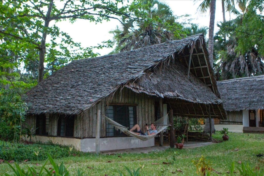 A person relaxes in a hammock on the porch of a rustic, thatched-roof cottage surrounded by lush greenery and palm trees, capturing the tranquil spirit of travelling through Africa—perhaps even along the serene coasts of Tanzania.
