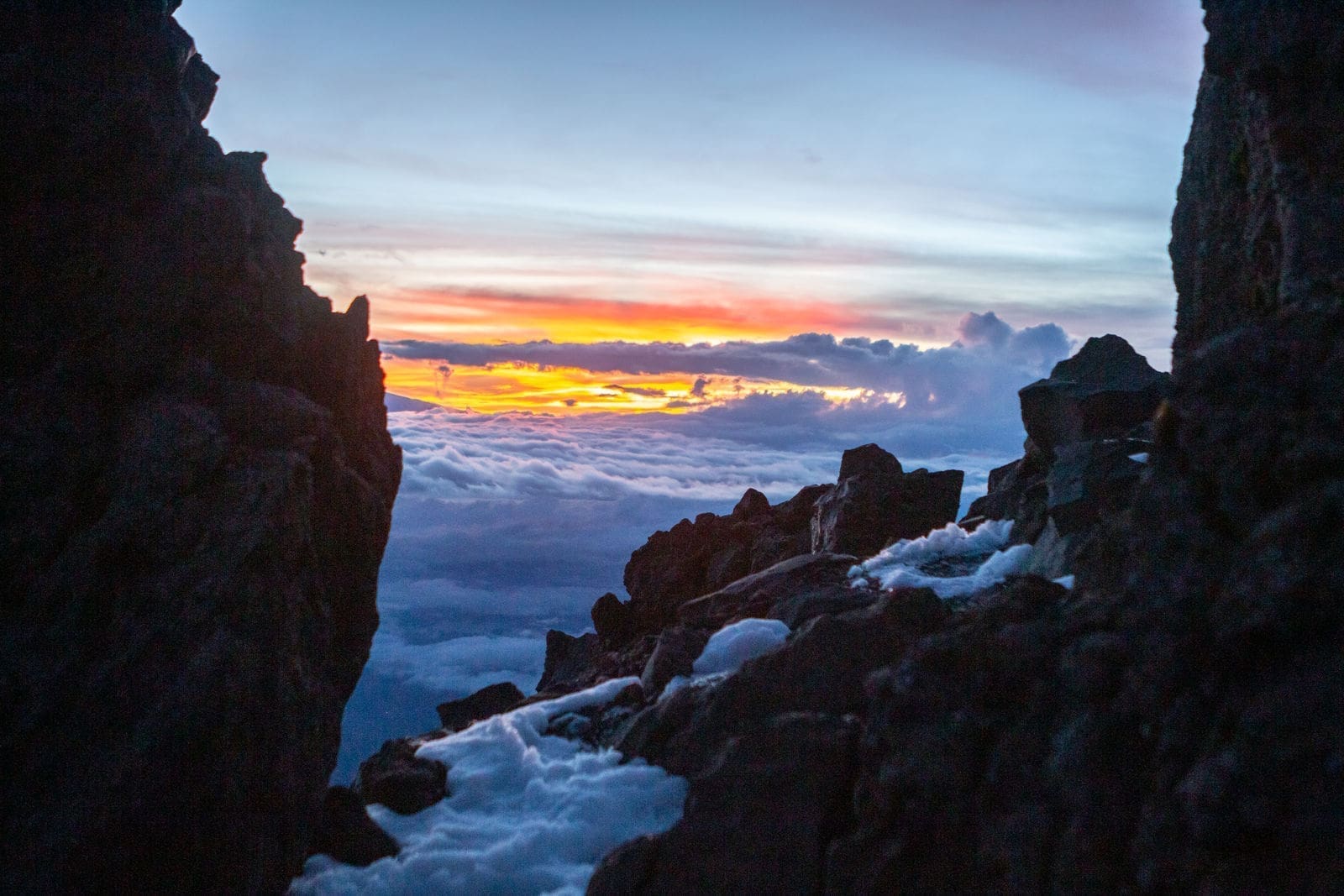 A vibrant sunset with orange and purple hues illuminates clouds below, framed by dark rocky cliffs and patches of snow—capturing the breathtaking beauty of travelling through Tanzania, Africa.