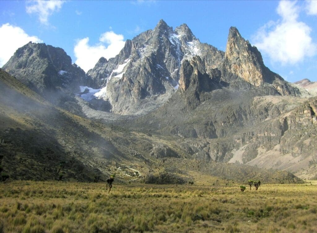 A dramatic view of Mount Kenya’s jagged peaks rising above grassy plains in Africa, with patches of snow and scattered clouds in the blue sky. The foreground features sparse vegetation and mist, enticing those travelling in search of adventure.