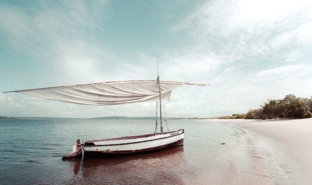 A small sailboat with a white sail is anchored at the shoreline on a calm, sandy beach in Mozambique. The sky is partly cloudy, and trees are visible in the distance—an inviting scene for travelling in Africa.