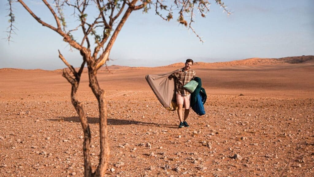 Eine Person in einem karierten Hemd und Shorts trägt eine Campingausrüstung, während sie durch eine felsige Wüstenlandschaft geht, mit einem einzelnen Baum im Vordergrund und sandigen Hügeln im Hintergrund.