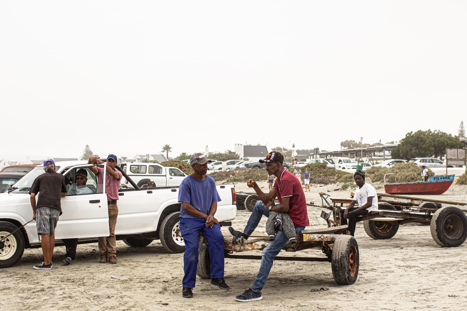 Several men gather on a sandy beach near white cars and makeshift carts. Two men sit and chat on a cart, while others stand or lean against a vehicle. A few buildings and trees are visible in the background.