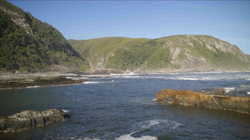 A coastal landscape in South Africa with blue ocean waves, a rocky shoreline, and green hills covered in vegetation under a clear blue sky—perfect for those who love travelling and exploring scenic vistas.