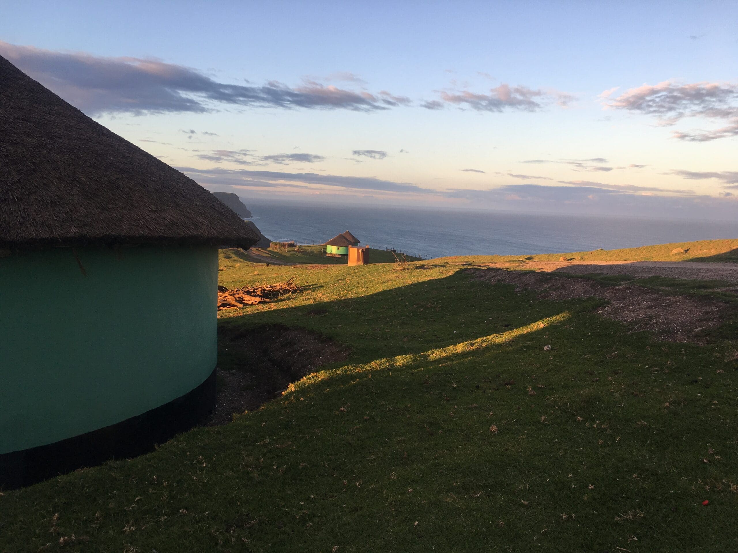 Een ronde hut met rieten dak staat op met gras begroeide heuvels die uitkijken over de oceaan, met meer hutten en een dramatische kustlijn zichtbaar in de verte onder een lucht met verspreide wolken.