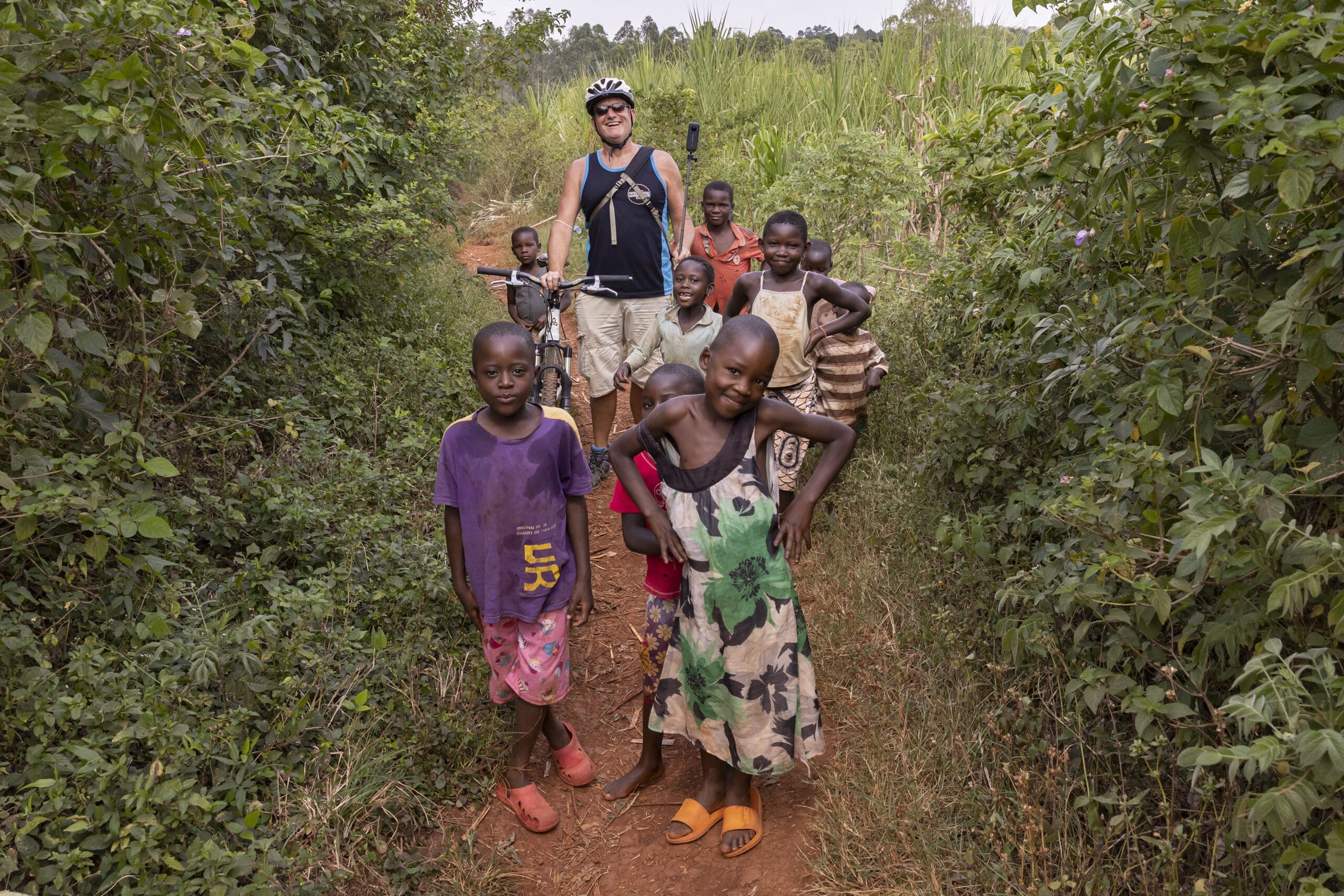 A group of smiling children stand on a dirt path surrounded by greenery, with a person wearing a helmet and sunglasses holding a bicycle and camera behind them. The children appear happy and playful.