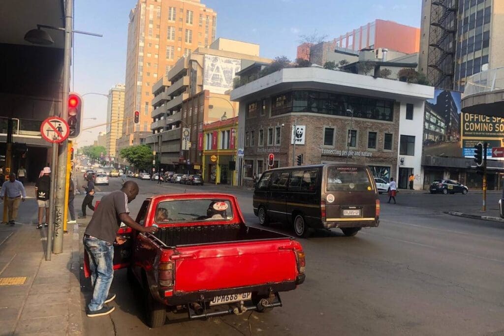 A man stands beside a red pickup truck parked on a city street corner. Tall buildings, traffic lights, and a van are visible in the background on a bright, sunny day.