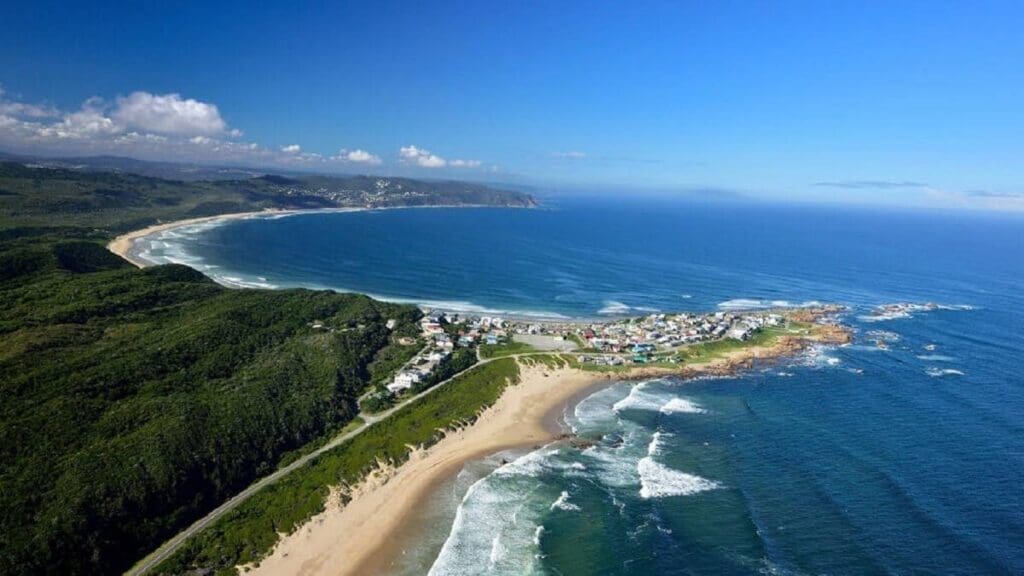 Aerial view of a coastal town in South Africa beside a curving sandy beach, lush green hills, and blue ocean waves under a clear sky with scattered clouds—a perfect scene for travelling enthusiasts.