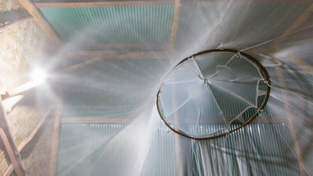 Nahaufnahme eines kreisrunden Moskitonetzes, das von der Decke hängt. Von der linken Seite des Bildes fällt Licht ein, das die Falten des Netzes und ein darüber liegendes Wellblechdach beleuchtet.