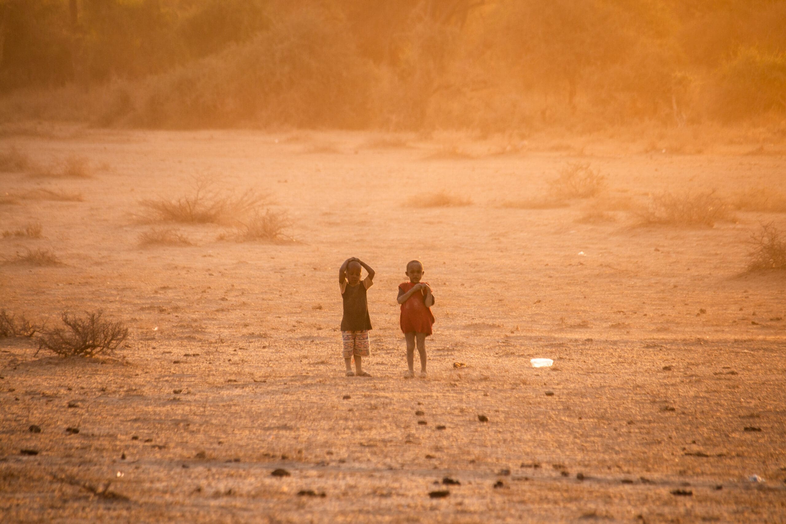 Zwei Kinder stehen auf trockenem, sandigem Boden mit verstreuten Büschen und schauen im goldenen Sonnenlicht von der Kamera weg. Die Landschaft wirkt karg und warm, und das weiche, dunstige Licht schafft eine ruhige Atmosphäre.