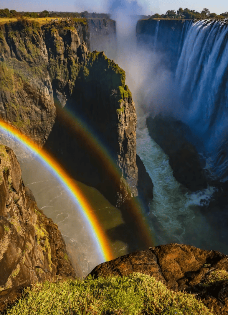 Een krachtige waterval stort zich in een diepe kloof, omringd door groene rotswanden. Twee schitterende regenbogen spannen zich uit boven de nevel die opstijgt uit het kolkende water beneden.
