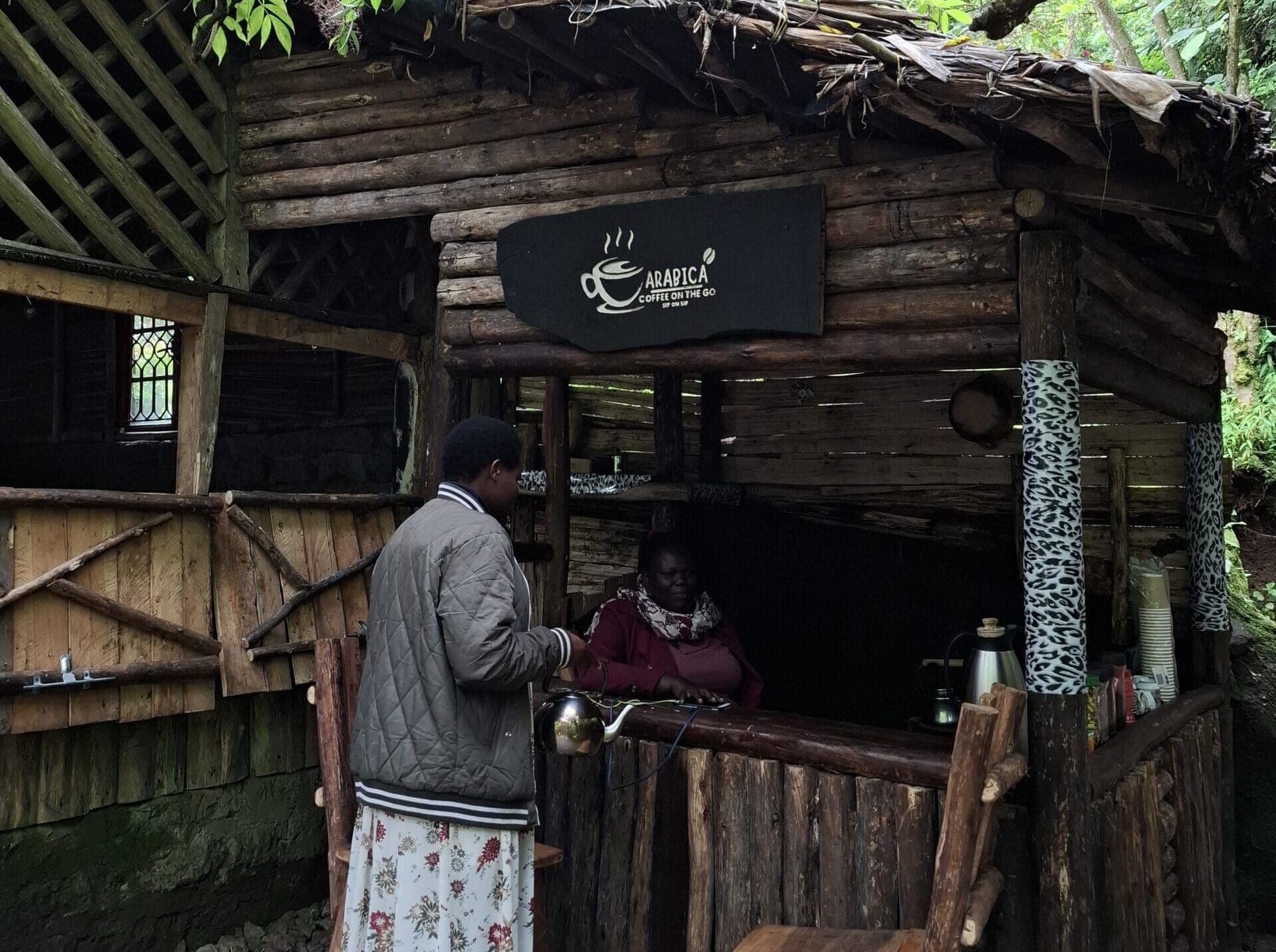 Een persoon staat bij een rustieke houten koffiekiosk met de naam Arabica Coffee Kiosk en heeft interactie met de verkoper binnen. De kiosk is populair bij reizigers die Afrika of Oeganda verkennen, wordt omringd door groen en heeft een gezellig, handgemaakt ontwerp.