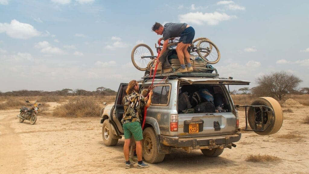 Two people secure a bicycle and luggage on top of an SUV parked on a dry, dusty road in Tanzania. The car’s rear door is open, and a motorcycle stands in the background amid sparse trees and the African desert landscape.
