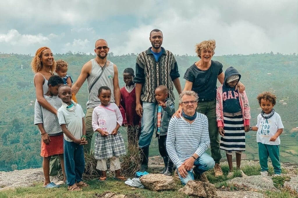 A group of adults and children stand and kneel together outdoors on a rocky area in Kenya, smiling for the camera with a scenic, green landscape and cloudy sky in the background—a joyful moment while travelling in Africa.