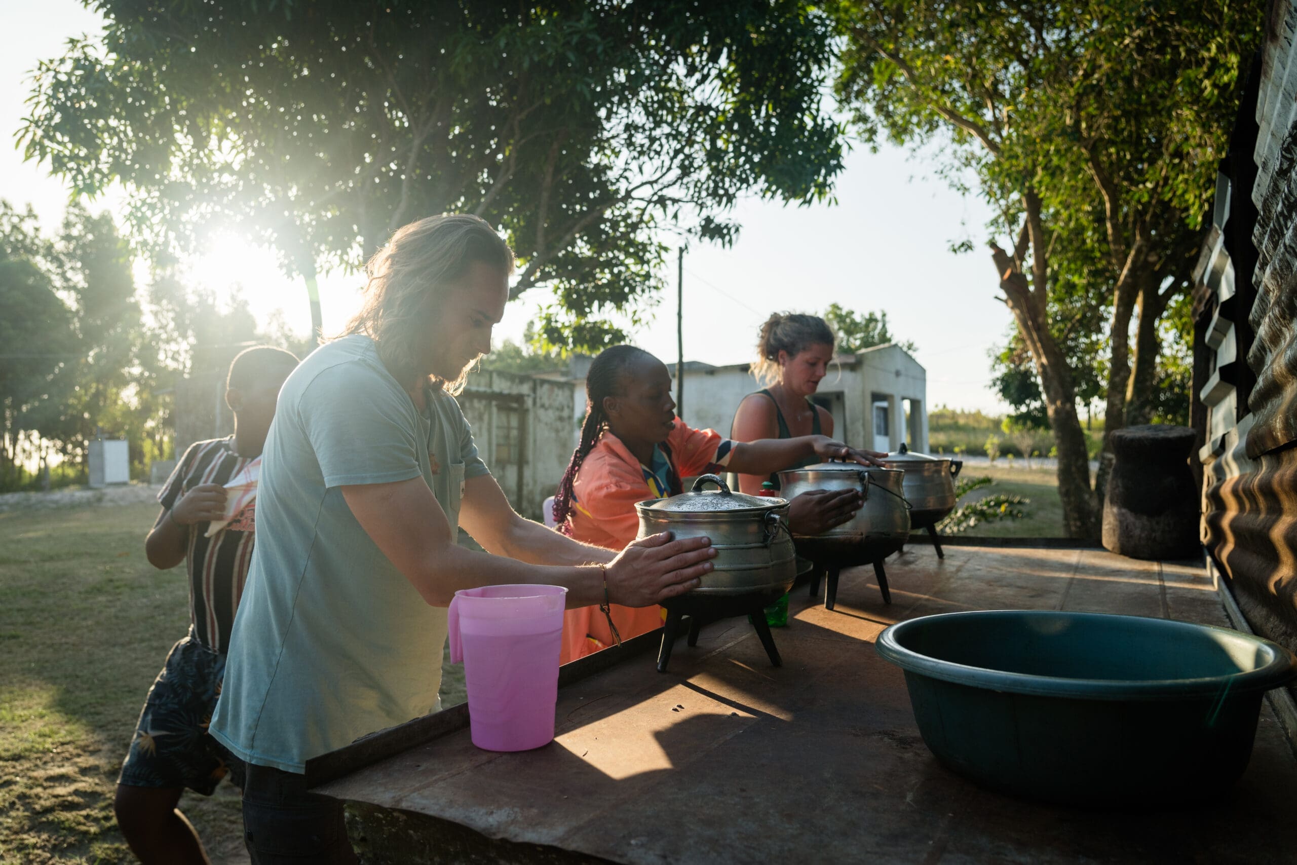 Three people stand outdoors at a long table, preparing food in pots under the sunlight. Trees and buildings are visible in the background, and a large green basin is on the table.