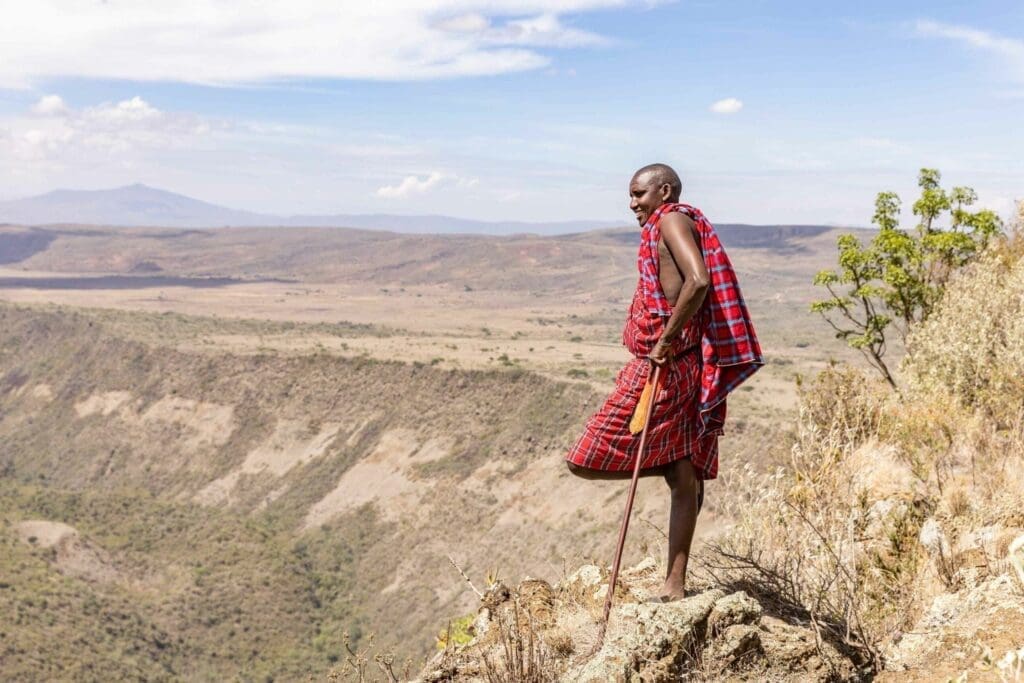 A Maasai man in traditional red clothing stands on a rocky cliff, holding a wooden staff, overlooking a vast, open landscape with hills and sparse vegetation under a partly cloudy sky.