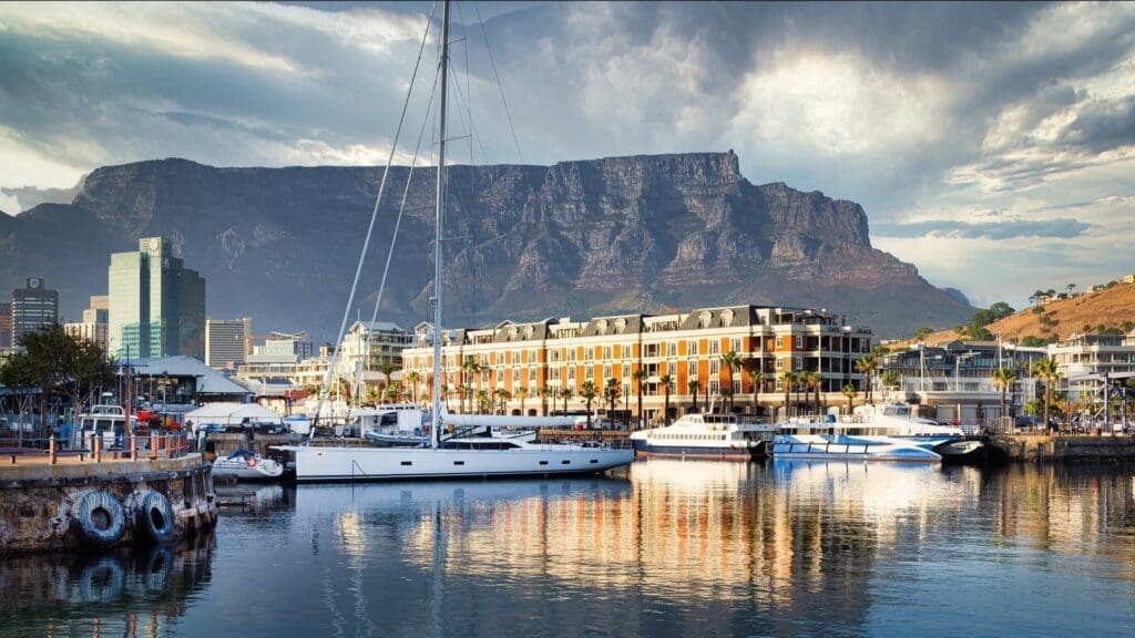 A marina with yachts and boats docked in calm water, stylish waterfront buildings, and the dramatic, flat-topped Table Mountain rising in the background under a partly cloudy sky—an iconic scene for anyone travelling in South Africa.