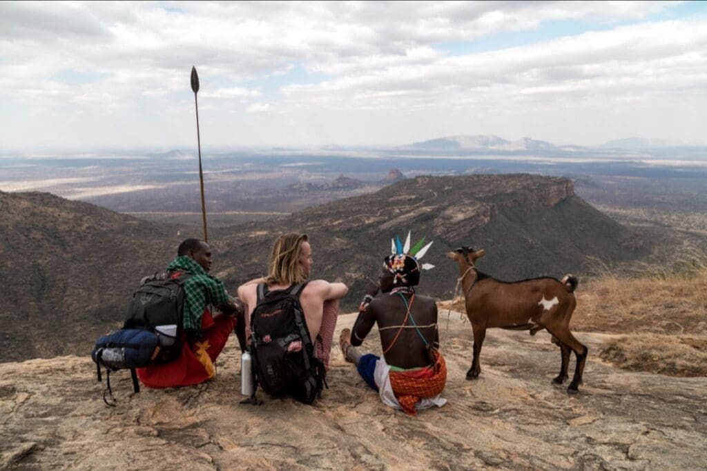 Three people—two in traditional attire with backpacks and one in casual clothing—sit on a rocky cliff overlooking a vast, hilly landscape, accompanied by a goat, under a cloudy sky.