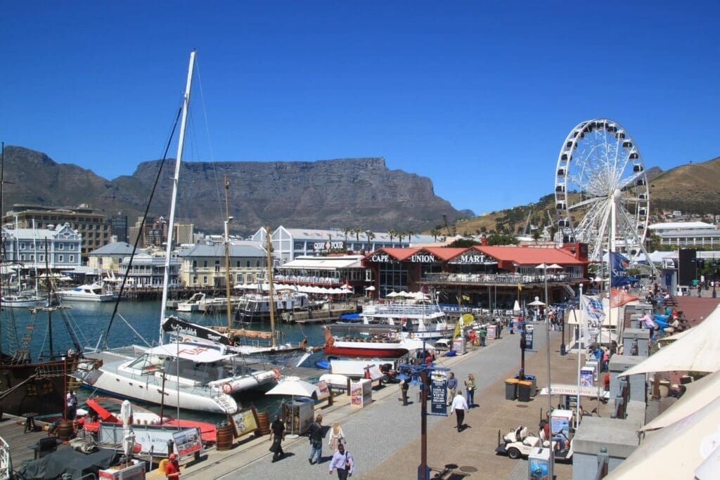 A busy waterfront scene in South Africa with boats docked at a marina, people walking, outdoor cafes, a large Ferris wheel, and Table Mountain in the background—a perfect spot for anyone travelling under a clear blue sky.