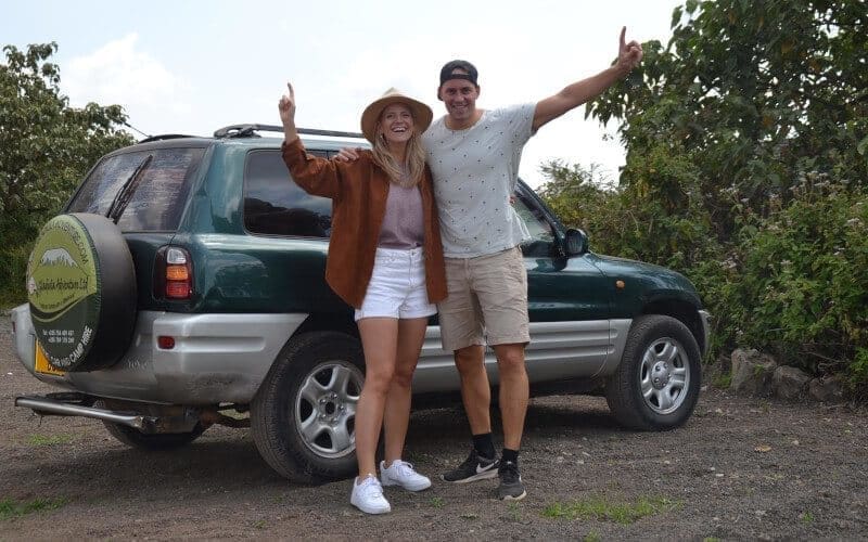A smiling man and woman stand beside a dark green SUV with their arms raised in excitement, enjoying travelling through Tanzania. They are dressed casually on a road trip, with lush green foliage and a partly cloudy African sky in the background.