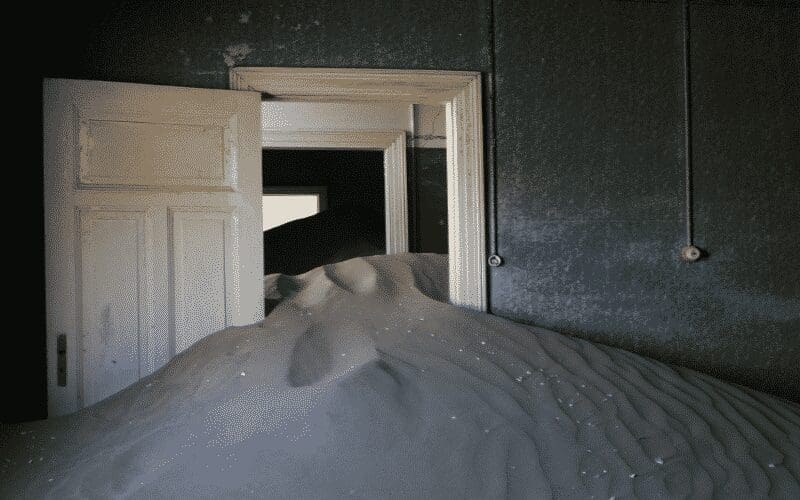 A room filled with sand nearly to the top of a white door, with more sand visible through a second doorway. The walls are dark, and the scene appears abandoned and overtaken by nature.