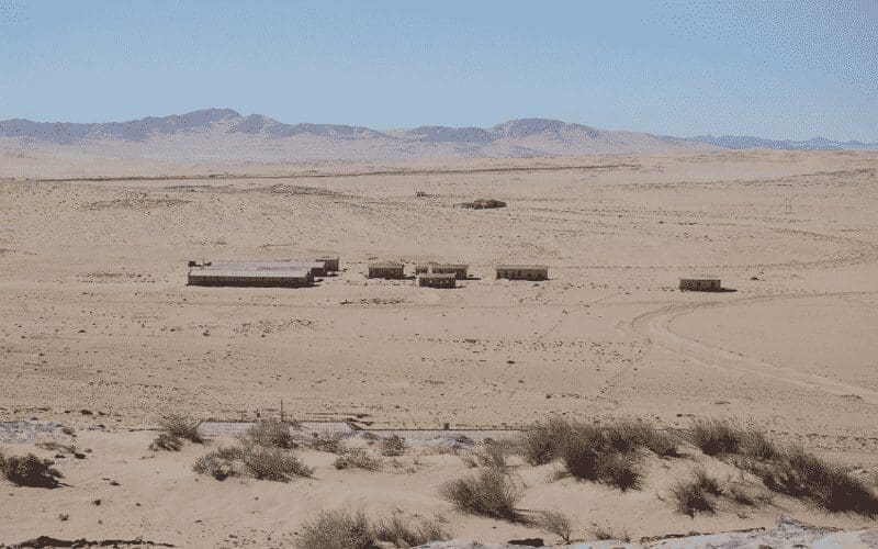 Een groep kleine, verweerde gebouwen staat geïsoleerd in een uitgestrekt woestijnlandschap onder een strakblauwe hemel, met bergen in de verte op de achtergrond en schaarse vegetatie op de voorgrond.