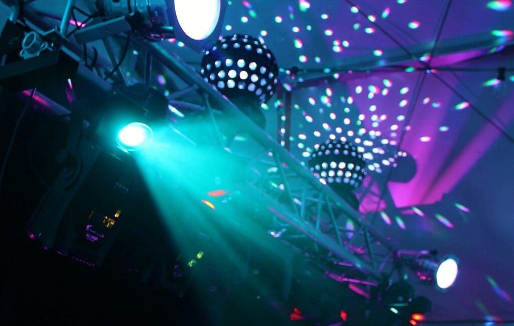 Bright stage lights and disco balls emit colorful beams and patterns onto a ceiling, creating a lively and festive atmosphere, likely at a concert or dance event. The scene is dominated by blue, purple, and teal lights.