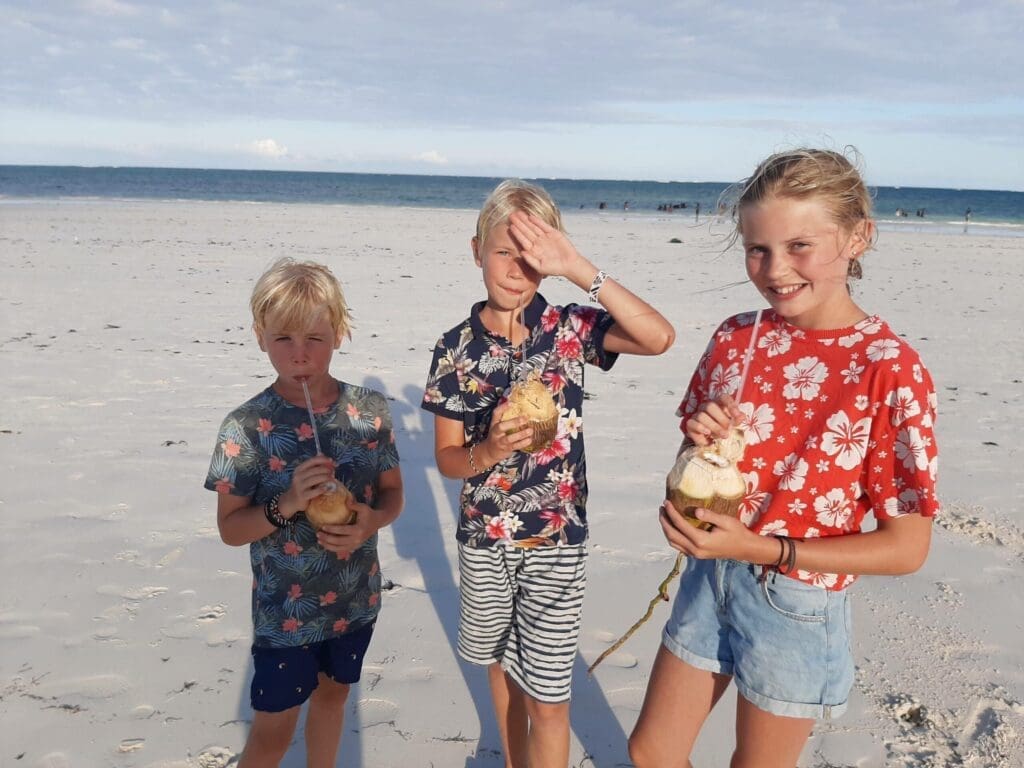 Three children stand on a sandy beach, each holding a coconut with a straw. They wear colorful, tropical-patterned clothes. The ocean and blue sky are visible in the background.