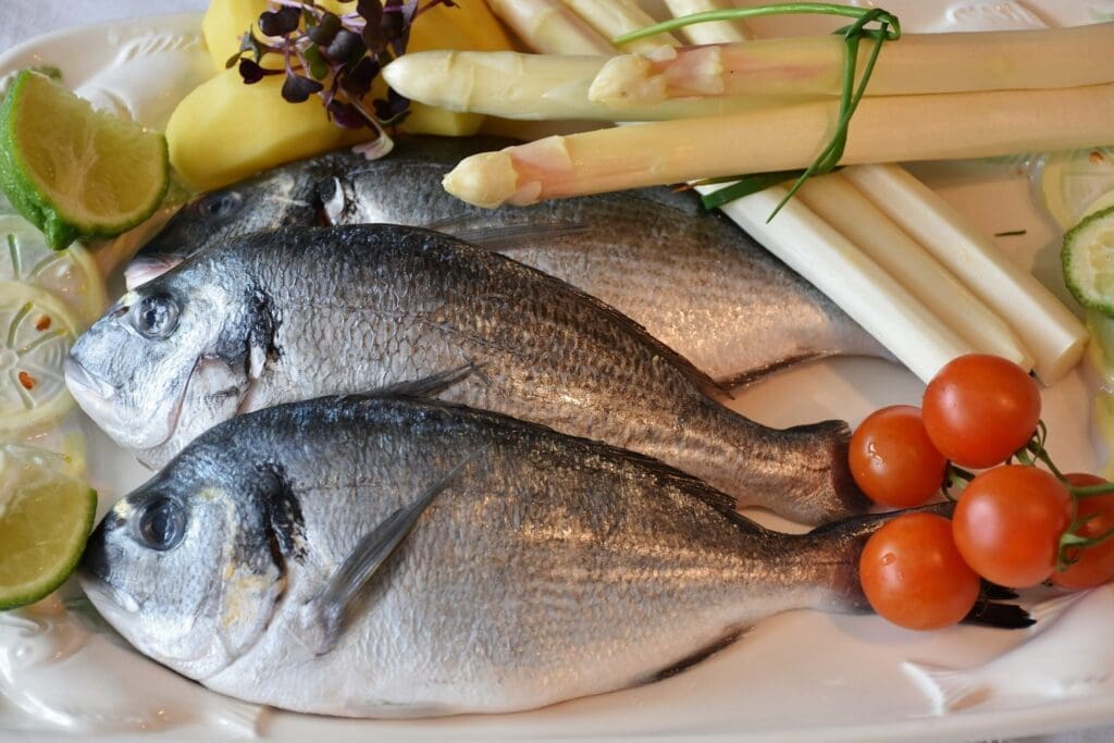 Three whole raw fish on a plate with fresh asparagus stalks, cherry tomatoes on the vine, lime slices, and ginger, arranged on a white rectangular platter.