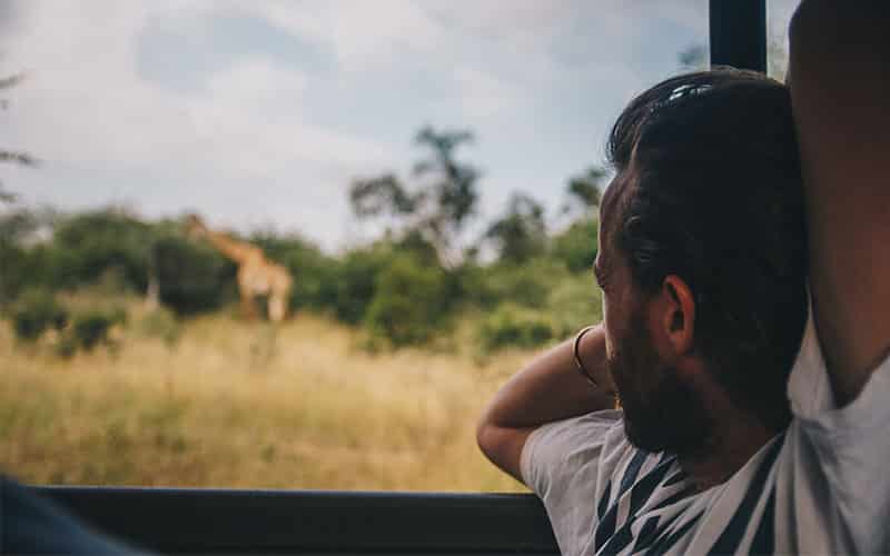 Een man zit in een voertuig, rust met zijn arm op het raamkozijn en kijkt naar een giraffe die in de verte tussen bomen en hoog gras staat. De scène is buiten, waarschijnlijk tijdens een safari.