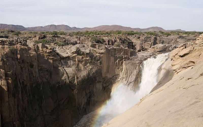 Een hoge waterval stort neer op een rotsachtige klif, met een regenboog aan de voet. Droog, ruig terrein en heuvels in de verte zijn zichtbaar onder een bleke, halfbewolkte hemel.