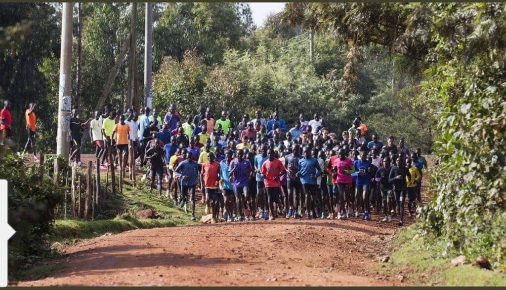 A large group of runners in colorful athletic wear gathers on a dirt road surrounded by lush greenery, preparing for a run in a rural outdoor setting.