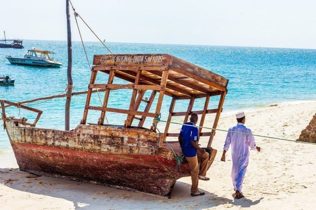Two men walk past an old wooden boat with a canopy on a sandy beach near the ocean; another boat floats in the water in the background under a clear, sunny sky.
