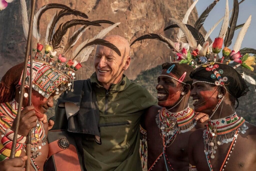 A smiling man in a green jacket stands with three people in colorful traditional attire, beaded jewelry, and feathered headdresses, travelling together outdoors in front of a rocky African landscape in Kenya.