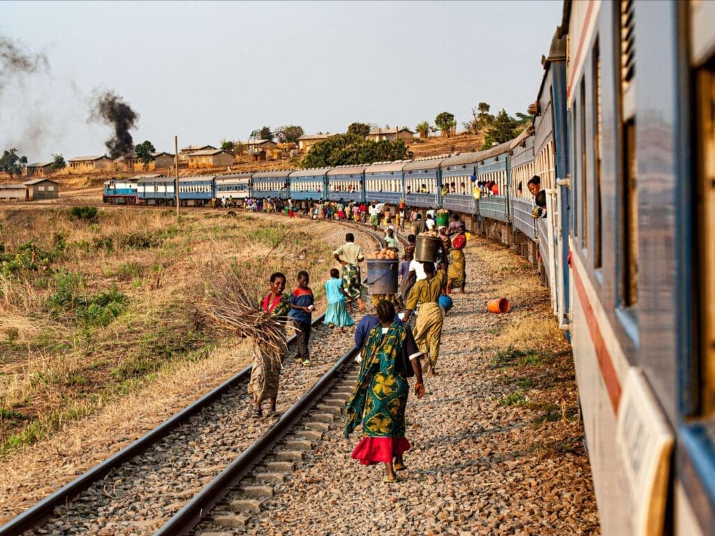People walk alongside a long, curving passenger train in a rural area, carrying bundles and containers. The landscape is dry, with scattered houses and another train visible in the background.