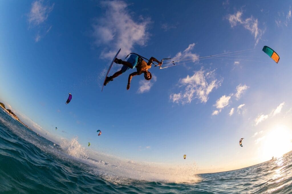 A person is performing an aerial trick while kiteboarding over the ocean, with several other kiteboarders in the background and a bright, clear sky overhead.