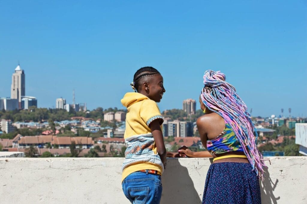 Two people stand on a rooftop, talking and looking over a cityscape with tall buildings under a clear blue sky. One has short hair in braids and wears a yellow hoodie, the other has long, colorful braids and a floral top.
