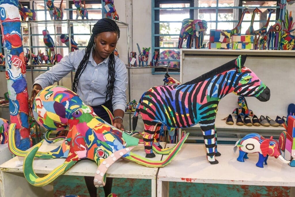 A woman arranges colorful animal sculptures, including a zebra, all made from recycled materials, on display shelves in a bright room with large windows.