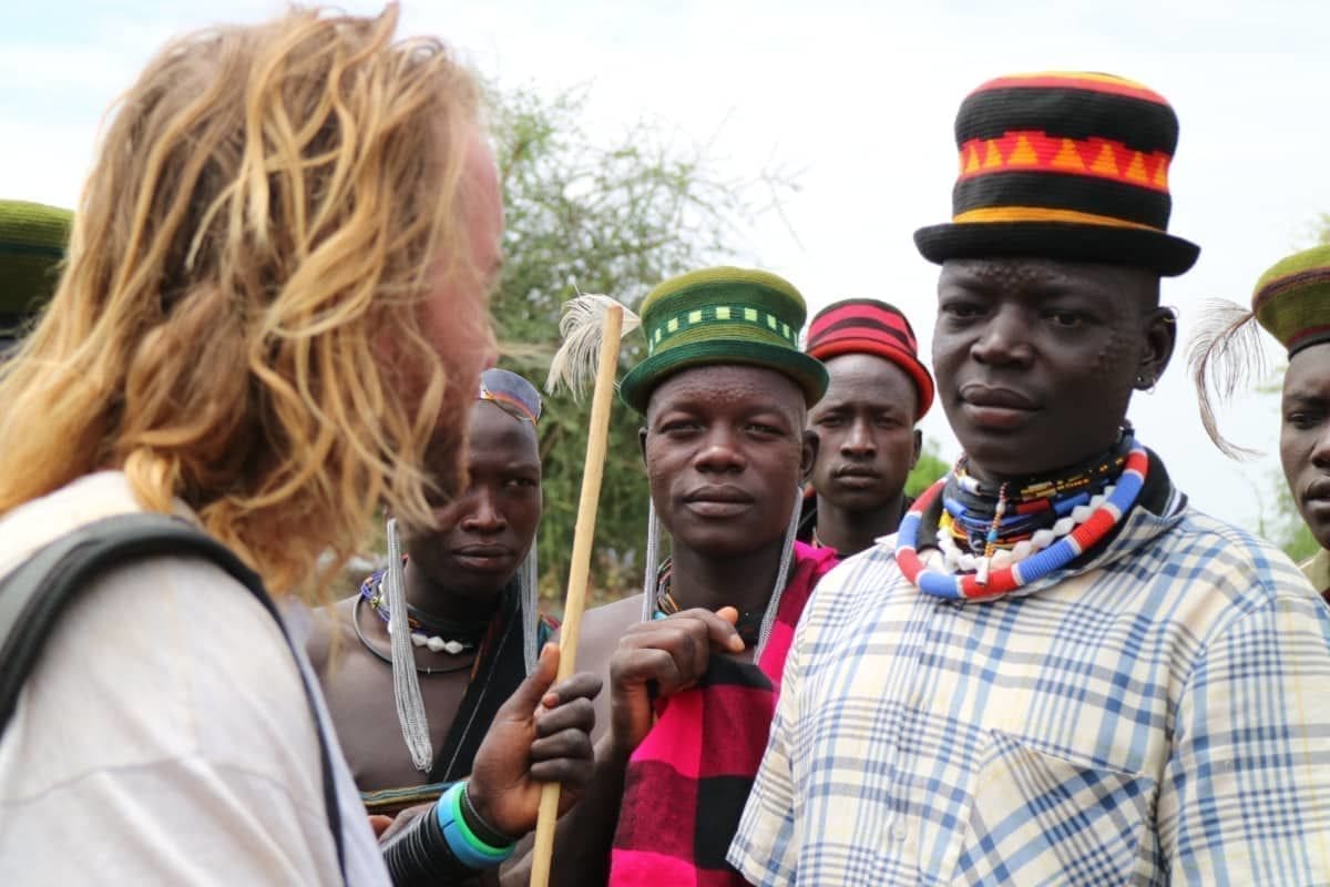 A man with long blonde hair speaks to three men wearing colorful hats and beaded necklaces, standing outdoors in Uganda with trees in the background. The group appears attentive and engaged in lively conversation while travelling through Africa.