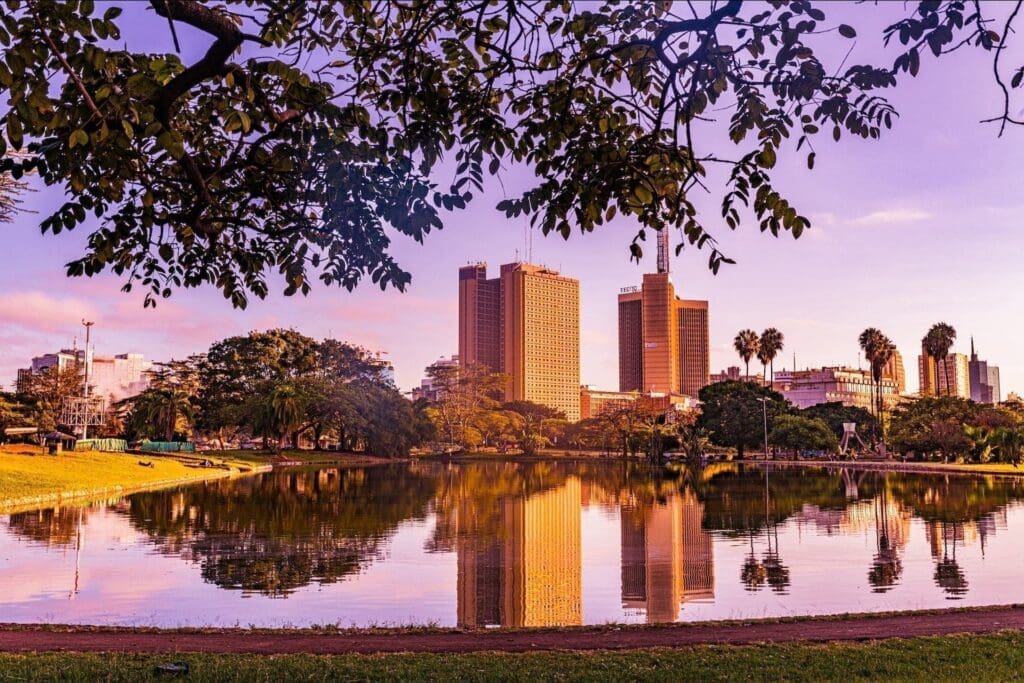 City skyline with tall buildings reflected in a calm lake at sunset, framed by tree branches and greenery in the foreground, under a purple and orange sky.
