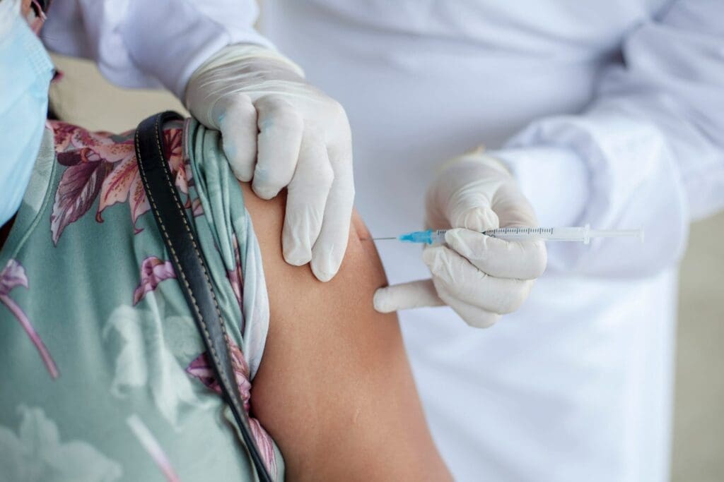 A healthcare worker wearing gloves administers a vaccine injection into a person’s upper arm. The person receiving the shot is wearing a floral shirt and a face mask.