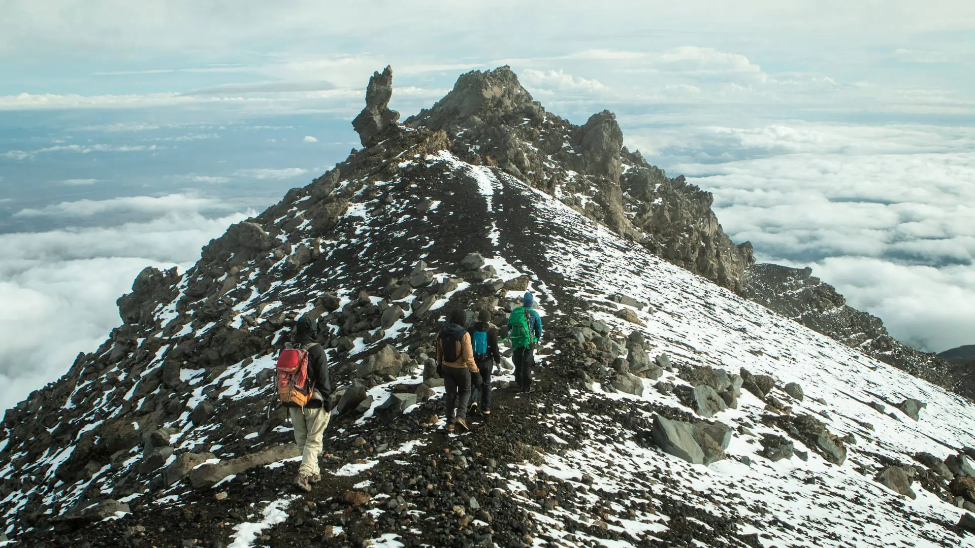 Vier Wanderer wandern entlang eines verschneiten, felsigen Bergrückens in Richtung eines zerklüfteten Berggipfels, umgeben von Wolken und einem dramatischen Himmel.