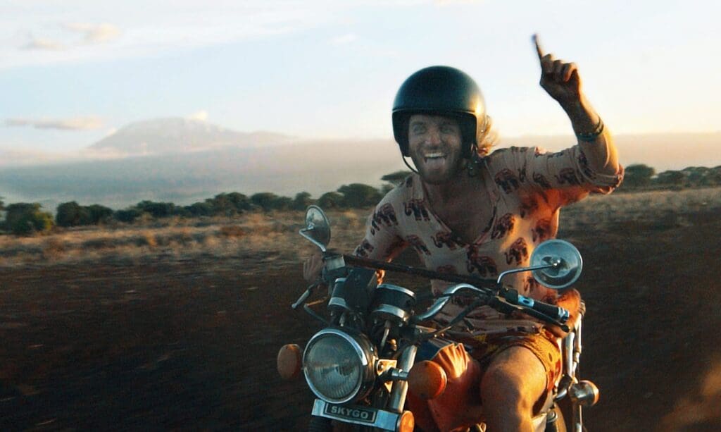 A person wearing a helmet and patterned shirt rides a motorcycle on a dirt road in Kenya, smiling and raising one hand with their index finger up. The landscape features open fields and distant mountains at sunset, capturing the spirit of travelling in Africa.