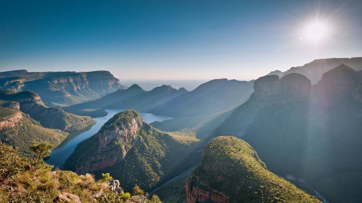Een schilderachtig uitzicht op een weelderig groene canyon in Zuid Afrika met kronkelende rivieren, ronde heuvels en dramatische kliffen onder een heldere hemel. Perfect voor reizigers die op zoek zijn naar avontuur, de zon werpt lange schaduwen over het adembenemende landschap.