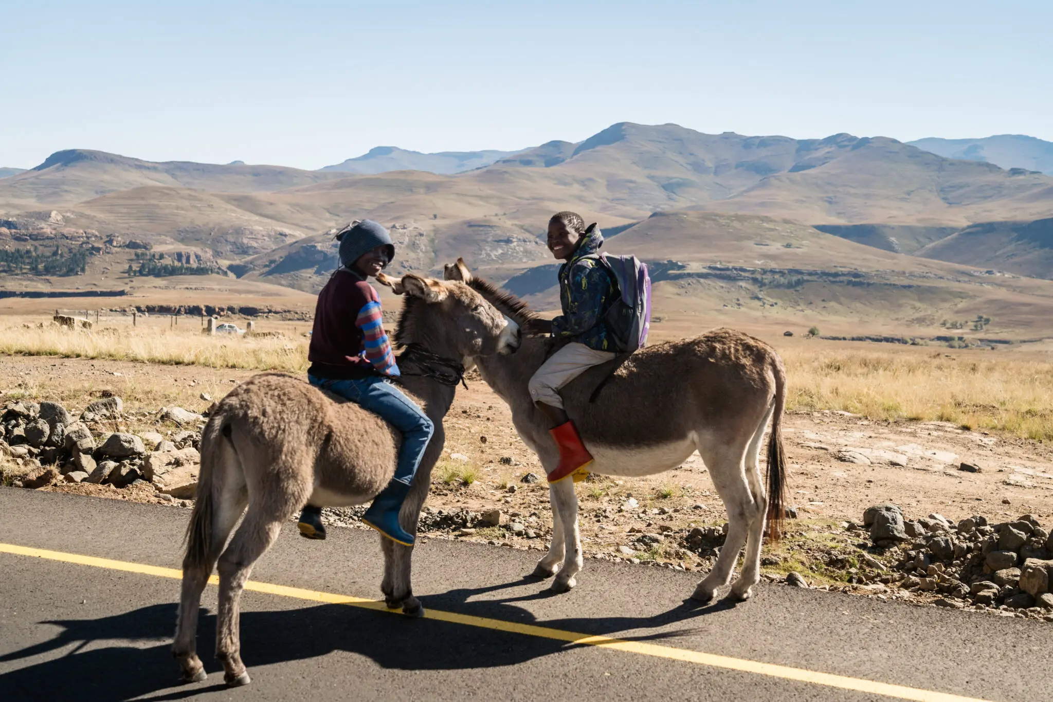 Zwei Menschen reiten auf Eseln am Rande einer Straße in einer ländlichen, bergigen Landschaft mit trockenem Gras, Felsen und Hügeln im Hintergrund unter einem klaren Himmel.