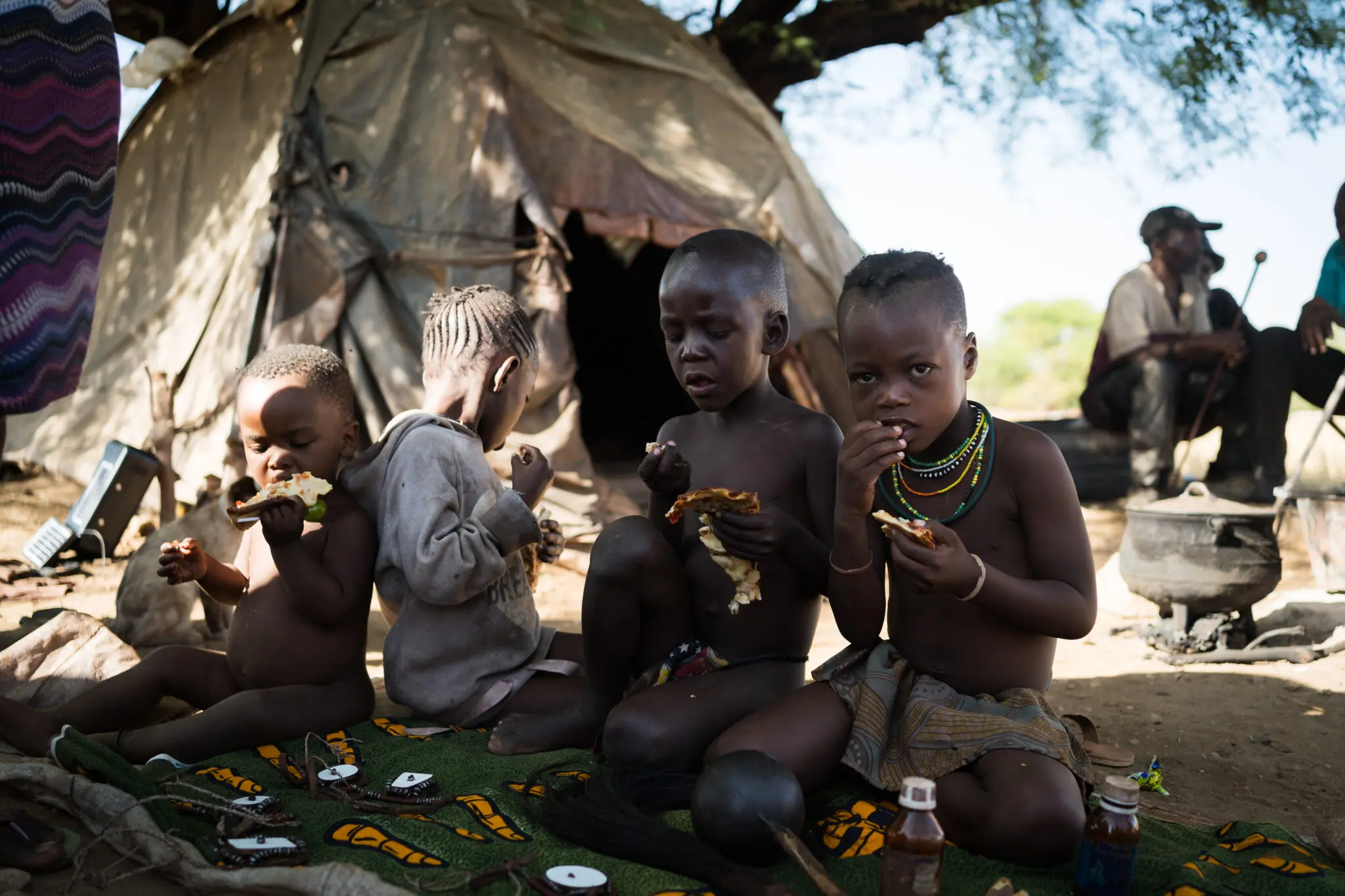Four young children sit on a patterned mat outside a makeshift tent, eating food. Two adults sit in the background near cooking pots and a tree, with sunlight filtering through. The setting appears rural and rustic.