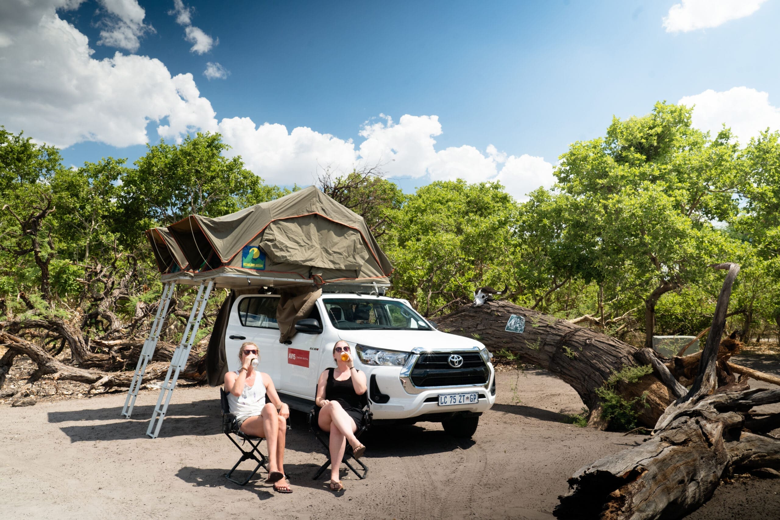 Twee mensen ontspannen met een drankje voor een witte Toyota pick-uptruck met een daktent, omringd door groene bomen en omgevallen boomstammen onder een gedeeltelijk bewolkte hemel - een beeld van de avontuurlijke geest van Botswana in Afrika.