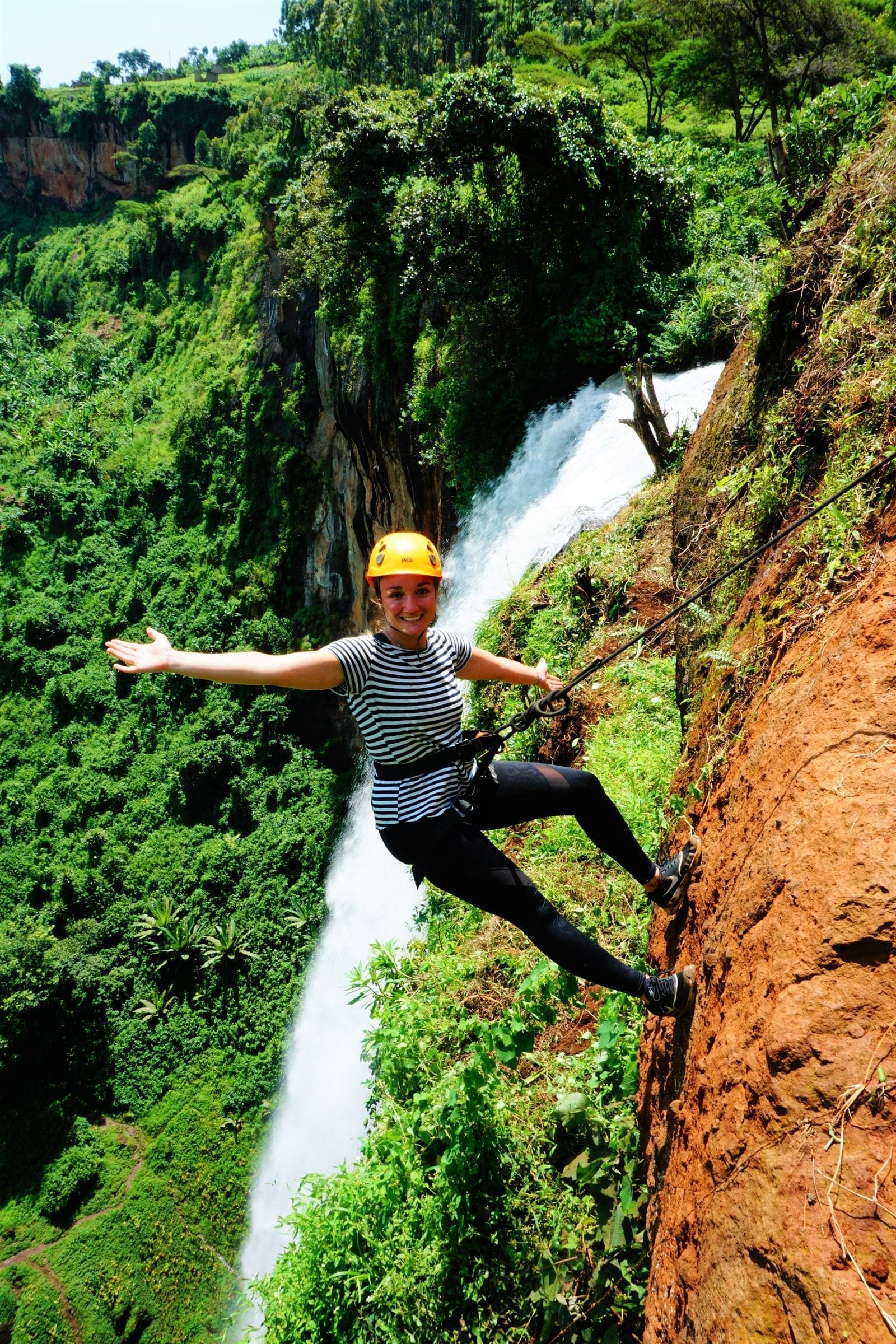 Een reiziger met een gestreept shirt, een zwarte broek en een helm abseilt van een steile, rotsachtige klif naast een hoge waterval in Oeganda, Afrika, omgeven door weelderige groene vegetatie, glimlacht en spreidt een arm wijd uit.