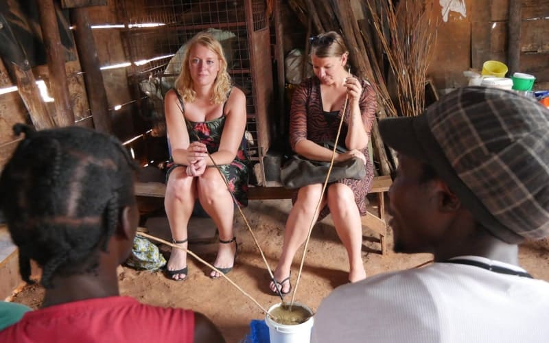 Vier reizigers zitten in een rustieke, schemerig verlichte kamer in Afrika; twee vrouwen kijken naar voren en twee anderen drinken met hun rug naar de camera uit een gedeelde emmer met lange rietjes.
